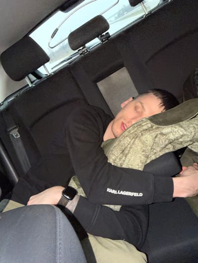 A male individual is depicted sleeping or resting in the rear passenger seat of a vehicle, in Gabarnitsa, Bulgaria. The subject, an adult male with short dark hair, has his eyes closed and mouth slightly ajar. He is wearing a black long-sleeved sweatshirt or hoodie with "KARL LAGERFELD" in white text visible on the left sleeve, and dark-colored trousers. A black smartwatch with a dark-textured band is present on his left wrist. His head rests on an olive green or camouflage-patterned fabric item, which also partially covers his lower body. The vehicle's interior features black fabric upholstery on the seats and headrests. Through the rear window, an indistinct outdoor scene is visible, suggesting a road or highway with guardrails and vegetation under an overcast sky. A silver-colored metal hanger-like object is suspended near the rear window.