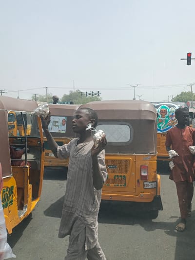 The image depicts a bustling street scene in Hotoro, Nigeria, on a clear, sunny day. In the foreground, a young male, dressed in a light-colored, striped tunic and trousers, walks towards the right. He holds small white bundles, enclosed in clear plastic bags, extended upwards in both hands, appearing to offer them for sale. Behind him and slightly to the right, a second male, wearing a reddish-brown tunic and trousers with sandals, walks towards the left, also holding similar white bundles in his left hand.
The individuals are positioned between several yellow and brown Keke Napep auto-rickshaws, which appear to be stationary or moving slowly in traffic. One prominent Keke Napep directly behind the foreground subject displays a visible license plate partially reading "SR-217M" and features a sticker with a smiling face on its rear panel. Further back, another Keke Napep is visible with distinctive green, black, and white artwork depicting a stylized skull or skeletal figure. In the background, an urban environment is present with power lines, utility poles, distant foliage, and a traffic light showing a red signal. The ground is asphalt.