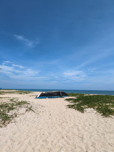 The image depicts a serene beach scene in Thuan An Ward, Vietnam. The foreground is dominated by a sandy beach dotted with scattered green vegetation. In the mid-ground, an overturned blue and black boat rests on the sand. The boat has some writing on its side, appearing to be "GHE + MA" followed by numbers. Beyond the beach, the calm blue ocean stretches to the horizon, meeting a clear blue sky with a few wispy white clouds. The overall impression is one of peacefulness and natural beauty, with no people visible in the frame.