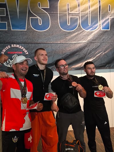 Four men are posing for a photograph in front of a banner that reads "EVS CUP SWISS ARM SPORT FEDERATION". All four men are wearing medals around their necks. The man on the far left is wearing a red and white shirt with a Swiss flag design and a white baseball cap. He is smiling and has his left arm raised. The man next to him is wearing a black t-shirt and orange pants. He is looking to the right and smiling. The third man from the left is wearing a black t-shirt and gray sweatpants. He is wearing glasses and holding a medal. The man on the far right is wearing a black t-shirt and black sweatpants. He is also wearing glasses and holding a medal. He is holding a red and white card with text on it. The event appears to be an arm wrestling competition, possibly held in Romanshorn, Switzerland, given the "SWISS ARM SPORT FEDERATION" banner.