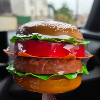 A close-up, low-angle shot captures a plastic hamburger toy, which appears to be a novelty item or air freshener. The toy is presented in its entirety, with the bun, lettuce, tomato, and a lower bun. The textures of the bun are detailed with embossed patterns resembling sesame seeds and the typical texture of baked bread. The lettuce is depicted as bright green, wavy plastic, and the tomato is a solid red disc. A clear plastic wrapper encases the entire toy. The toy is positioned in a car interior, with the dashboard and dark interior visible in the blurred background. Hints of an outdoor environment with buildings and greenery are also discernible in the background, suggesting the car is parked or in motion. The lighting is natural and diffused, with a slight overcast sky indicated by the soft illumination. The image does not depict the city of Ravnogor, Bulgaria.