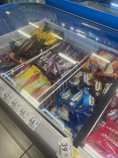 The image shows a freezer chest filled with various ice cream products. The ice cream packages are mostly in wrappers with Cyrillic text. There are several types of ice cream visible, including some in gold wrappers, some in purple wrappers with images of berries, some in orange wrappers, and others in brown and blue wrappers. A price tag with the number "46" and other symbols is visible on the front of the freezer. The freezer has a glass lid, through which the products are viewed. The overall environment appears to be a retail setting, likely a supermarket or convenience store. The location is identified as Гожули, Ukraine.