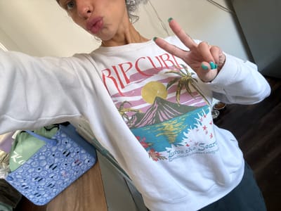 A woman is posing for a selfie, wearing a white sweatshirt with "RIP CURL" printed on the front with a beach scene. She is making a peace sign with her left hand, which has green nail polish. Her face is visible, showing her nose and lips. A blue plastic basket with a white seashell pattern is in the background, as well as a wooden table and a wall. The location in the city of Seal Beach, United States of America, cannot be determined from the available information.