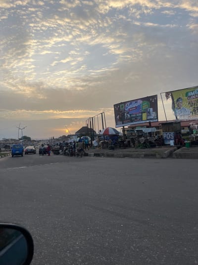 The image captures a street scene in Offatedo, Nigeria. The sky is filled with clouds, illuminated by the setting sun. The road, occupying the right side of the frame, appears to be paved with asphalt. On the left side, a busy commercial area is evident, featuring large billboards advertising "Sallah Promo" with a "50% DISCOUNT" and "POS" services. Additionally, there is street vending and groups of people and motorbikes. In the distance, the sun is setting.