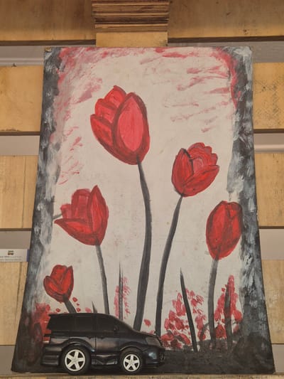 The image shows a painting of red tulip-like flowers with black stems, on a surface that appears to be a piece of repurposed wood or board. The painting has a grey and white border with some red smudges. A black toy car is placed on the right side of the painting, partially obscuring the flowers. The painting is resting on a wooden pallet. A partial label indicating "BUS TRIPS to and from Cubao, Bacolod, Manila" is visible near the bottom of the image, suggesting a possible location in the Philippines. Given the reference to Cubao, Bacolod, and Manila, it may be located within Pasay, Philippines.