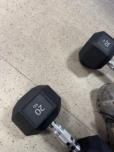 The image presents a high-angle view of a section of a gym floor. Two 20kg black dumbbells rest on the speckled, light-grey flooring. A portion of a grey athletic shoe, possibly belonging to a person not fully visible in the frame, is positioned near one of the dumbbells. The dumbbells feature hexagonal ends and knurled metal handles. The flooring is clean. The setting suggests a workout area. No further details indicate the city of Babergh, United Kingdom.