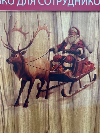 In Kadamjay, Kyrgyzstan, a decorative panel features a depiction of Santa Claus riding in a sleigh pulled by a reindeer. The panel is made of wood with a visible grain texture. Santa Claus, wearing his traditional red suit and hat, is seated in a dark-colored wooden sleigh laden with red gift boxes. The reindeer, with large antlers, is positioned to the left of Santa, pulling the sleigh. The background of the image is a light brown wood grain. At the top of the image, a red banner with light blue Cyrillic lettering is visible. The scene appears to be an outdoor decoration or advertisement, likely related to a winter holiday theme.