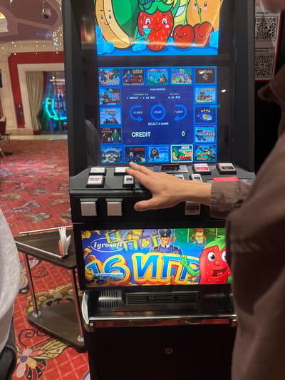 The image shows a person's hand interacting with a slot machine in what appears to be a casino or entertainment venue. The slot machine has a bright, colorful display featuring cartoon characters and game titles such as "Crazy Monkey," "Sweet Life," and "Fruit Cocktail." The denomination is set to "1 CREDIT = 1.00 RUB," and the balance is "0.00 RUB." The person's hand is positioned over the buttons, suggesting they are playing the game. In the background, through a glass partition, other slot machines with neon lights are visible, along with a red carpet with intricate patterns. The environment suggests an indoor entertainment setting. No specific details about the city of Shkotovo, Russia, are present within the image itself.