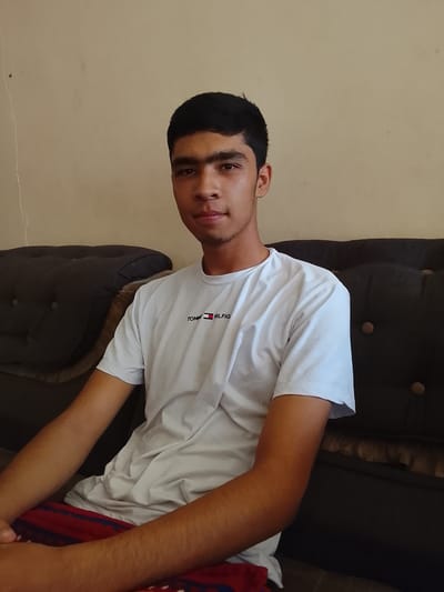 A young man with dark hair and brown eyes is seated on a dark sofa in what appears to be a home environment. He is wearing a white t-shirt with the Tommy Hilfiger logo and red and blue striped pants. The wall behind him is a neutral beige color, and there is a thin crack running vertically down the wall. The lighting suggests it is daytime, with natural light coming from the side. The image is a medium shot, focusing on the upper body of the man. The location of Kasri Arifan, Uzbekistan, is noted as the setting for this scene.
