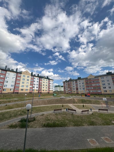 The image shows a residential area in Ashmyany, Belarus, on a partly cloudy day. In the foreground, there is a paved walkway made of grey brick pavers, with a lamppost on a metal pole to the left and two circular manhole covers embedded in the pavement. Beyond the walkway, there is a grassy slope with several paved pathways winding through it. Benches are placed along these pathways. In the middle ground, there are two large apartment buildings, one on the left and one on the right. The building on the left is multi-story and features a facade with sections of white, yellow, and red brick. The building on the right is similar in height and color scheme, with grey and red brick elements. A playground with colorful equipment is visible in front of the apartment buildings. Further in the background, more apartment buildings with green roofs can be seen, indicating a developed residential neighborhood. The sky is a vibrant blue, populated with fluffy white clouds. The overall impression is that of a modern, well-maintained urban environment.