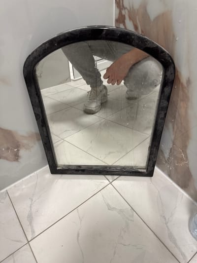 The image shows a person squatting in front of a mirror. The person is wearing grey sweatpants and white sneakers. Their left hand is visible, resting on their knee. The mirror has a black frame and is positioned in a corner where two walls meet. The walls have a marbled pattern and appear to be tiled. The floor is also tiled with a white marble-like pattern and shows signs of wetness or residue. The overall scene suggests an indoor environment, possibly a bathroom or a room undergoing renovation, with the mirror leaning against the wall. The location of Madan, Bulgaria, is an external detail not depicted within the image itself.