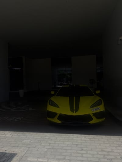 The image depicts a yellow sports car parked in a dimly lit indoor space, likely a parking garage or entrance area. The car has a black stripe running down the center of its hood and a license plate reading "Dubai X 69750". The car is positioned next to a sidewalk paved with grey bricks, and there's a painted "G4" marking on the ground next to it. The background reveals a building with glass windows or doors, and the overall scene suggests an urban environment, possibly in the Business Bay area of the United Arab Emirates. The left side of the image is much darker, showing a wall and ceiling.