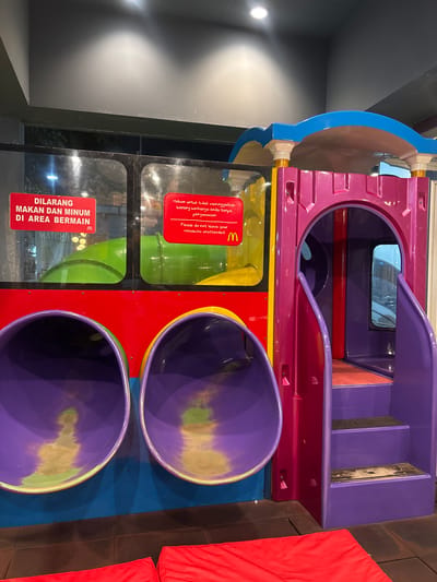 The image shows a McDonald's indoor playground structure in Bencongan Indah, Indonesia. The structure has a pink and purple train-like top section with a small window and a set of stairs leading up. Below, there are two circular openings in a red and blue section, with purple rims. Warning signs are affixed to the structure in Bahasa Indonesian and English; they forbid eating and drinking in the playground area and warn people not to leave valuables unattended. Green glass panes are visible near the warning signs. Red pads are visible near the base of the structure.
