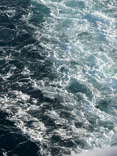 The image displays a detailed, overhead perspective of turbulent seawater within the marine environment surrounding Kamariotissa, Greece. The water surface is heavily agitated, characterized by an extensive distribution of white foam and aerated water, indicating significant churning. The water's coloration ranges from deep dark blue and greenish-blue in calmer or deeper sections to lighter shades of turquoise and white where foam is most concentrated and breaking. The foam forms intricate, irregular patterns across the entire visible surface. No individuals, vessels, or identifiable man-made objects are present within the frame. The scene captures a dynamic aquatic environment, focusing exclusively on the texture, movement, and color variations of the agitated water.