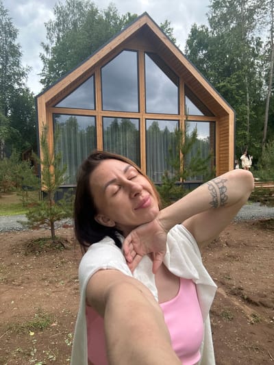 In this photograph taken in Severnyy, Russia, a woman with her eyes closed is positioned on the right side of the frame, her head tilted back and her hand near her face. She is wearing a pink top and a light-colored wrap, partially covering her. A tattoo is visible on her arm. Behind her, a modern wooden cabin with large, dark-paned windows dominates the left side of the image. The cabin has a distinctive A-frame design. The surrounding area is natural, with trees and ground covered in dirt and sparse vegetation. Two other figures, appearing as small silhouettes in the distance on the left, are standing near the cabin. The overall impression is one of relaxation and leisure in an outdoor, possibly rural or semi-rural, setting. The lighting suggests it could be daytime, possibly overcast given the muted tones and reflections in the windows.