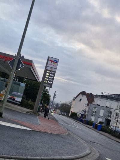 An outdoor, slightly tilted photograph captures a street scene in Niedernhausen, Germany, under an overcast sky. In the foreground on the left, a tall grey utility pole stands, featuring a black and white diamond-shaped sign displaying the number "5". To its right, a portion of a building with "Energie" visible on its facade indicates a TotalEnergies gas station. A prominent, slender digital totem displaying the TotalEnergies logo and fuel prices, including "ESSAI 2.23€", "2.40€", "2.09€", and "2.32€ Excellium", is located in the midground.
One individual, dressed in a dark jacket over a lighter garment and carrying a dark bag, walks on a red-brick patterned sidewalk adjacent to the gas station totem. The asphalt road curves slightly downhill towards the background on the right. Along the right side of the street, residential buildings are visible, including a light-colored house with a red-tiled roof and a grey multi-story building with white roller shutters on its windows. Several blue and yellow recycling bins are positioned along the curb in front of the residential properties. Distant streetlights or vehicle headlights are discernible further down the road. The sky is largely grey and cloudy, indicative of an overcast day.