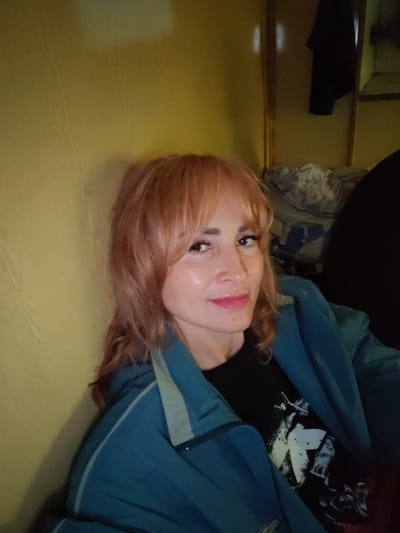 A medium shot captures a woman with auburn hair and bangs, wearing a teal jacket over a black t-shirt, leaning against a yellow-toned wall. Her head is tilted slightly, and she is looking towards the camera with a gentle smile. Her makeup includes lipstick, eyeliner, and mascara. The background shows a dark, indistinct area with some folded fabric and a dark garment hanging on a wall. The lighting is warm and somewhat dim, creating shadows. The scene appears to be indoors, possibly a room or a dimly lit space. There is no indication of the city of Andrukovo, Russia, or any specific urban environment.