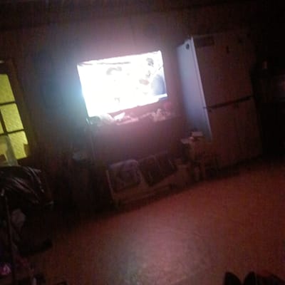 The image shows a dimly lit room with a television screen displaying an image. To the left of the television is a window with a yellow tint. In front of the television, there are some objects on a surface, and below that are two dark rectangular objects. To the right of the television, there is a tall, light-colored appliance, possibly a refrigerator or a cabinet. The floor is a reddish-brown color. The overall scene appears to be a domestic living space. No specific location within Prikubanskiy, Russia, can be determined from the image.