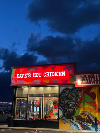 The image shows a Dave's Hot Chicken restaurant in East Brunswick Township, United States of America. The building has a large window facade and is adorned with a colorful mural and the restaurant's neon sign. The sky is dark blue, indicating that it's either evening or early morning. A vehicle is parked on the side of the building.