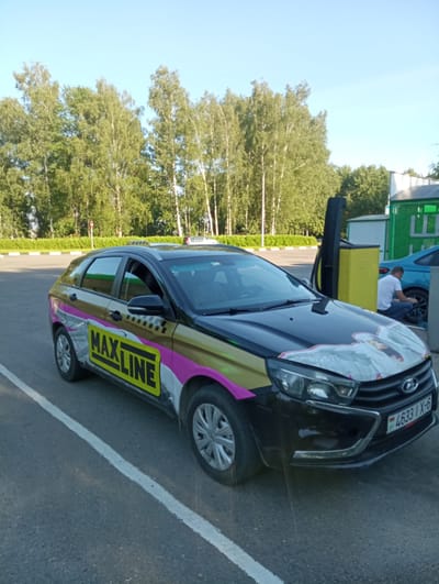 A black Lada Vesta hatchback, branded with "MAXLINE" graphics and an image of a swan on its hood, is parked on an asphalt surface next to a white line, possibly in a parking lot. The car has a taxi light on its roof. In the background, a person in a white t-shirt and blue jeans is kneeling by the tire of a blue car, near a yellow charging station. A line of birch and other trees with green foliage borders the scene under a clear blue sky. A license plate on the Lada reads "4633 IX-6". The scene appears to be set in the vicinity of Буйніцкі сельскі Савет, Belarus.