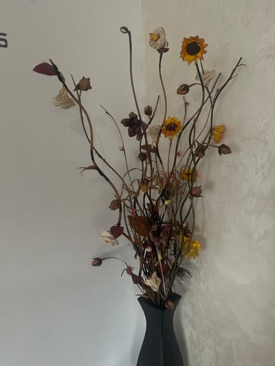 A still-life photograph features a bouquet of dried flowers and foliage in a dark-colored, angular vase. The bouquet includes various types of flowers, some resembling sunflowers and roses, in shades of yellow, white, and brown. The stems and branches are brown and twisted, suggesting a natural, dried arrangement. The vase is positioned on a light-colored surface, and the background appears to be a light-colored wall. The overall composition has a somewhat rustic or vintage aesthetic. The photograph does not depict Stroevo, Bulgaria, nor does it include people, actions or interactions.
