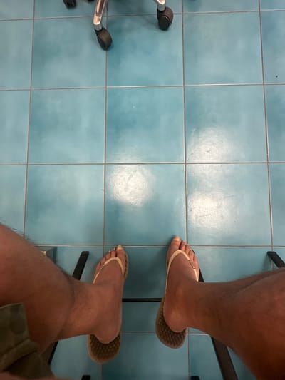 A downward-facing, first-person view captures the lower legs and feet of an individual seated in Kfar Vitkin, Israel. The subject's bare, tanned, and hairy legs are visible, with the left leg partially obscured by a garment. Both feet are adorned with light-colored, possibly beige, flip-flops featuring brown, textured soles. The toenails display a light, possibly yellowish or beige, polish. The feet are resting on a dark, indistinct object, possibly chair legs or a footrest. The immediate environment consists of a floor covered in square, light blue or teal ceramic tiles with dark gray grout lines. The tiles exhibit minor scuff marks and reflections of overhead illumination. In the upper portion of the frame, the chrome base and black caster wheels of an office chair are visible, also resting on the tiled floor.