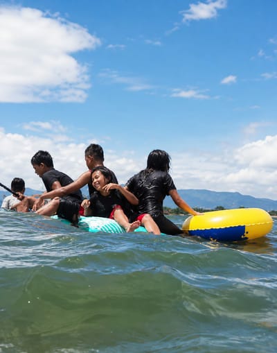 Six individuals, appearing to be children and possibly one adult, are on a makeshift raft in the water. They are all wearing black tops. The raft is constructed from a surf mat and a large inflatable ring. The inflatable ring is yellow and blue. The water is green and choppy, with ripples and waves. In the background, there are mountains, and the sky is blue with scattered white clouds. The scene appears to be a recreational activity taking place in the open water. The location is identified as Lumban Bul Bul, Indonesia.