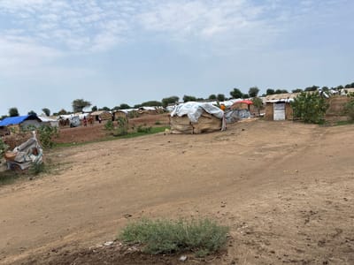 The image depicts a refugee camp in Ad-Damazin, Sudan, characterized by numerous temporary shelters constructed from natural materials like straw and covered with tarps. The shelters are densely packed, with pathways made of dirt and gravel running between them. A few individuals are visible in the distance, interacting within the camp. The environment is arid, with sparse vegetation and a cloudy sky. The overall scene suggests a makeshift and basic living environment for the camp's inhabitants.