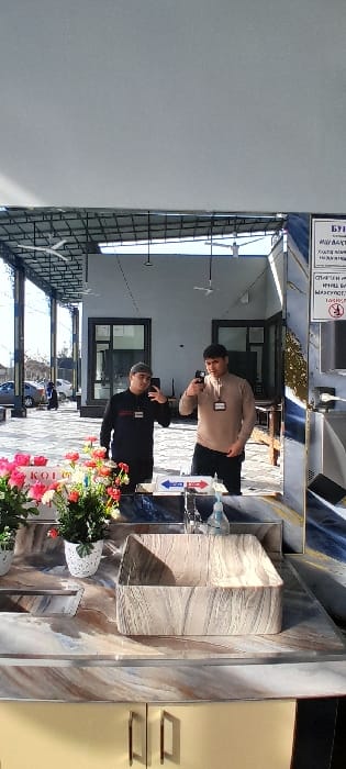 The image shows the reflection of two men in a mirror. One is wearing a black top and a baseball cap, and the other is in a light-colored top. They are both looking at the camera and seem to be taking a selfie. In front of them is a floral arrangement with a sign. The background includes an outdoor area with vehicles and a building with windows and overhead fans. The reflection also shows a desk, a patterned wall, and light yellow cabinets.
