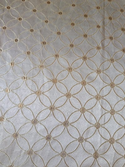 The image displays a close-up, overhead view of a textured fabric with a repeating pattern of interlinked circles. The fabric appears to be a light, neutral color, possibly cream or pale gold, and has a subtle sheen. The pattern is created by thin, embroidered lines in a slightly darker shade, forming a grid of overlapping oval shapes. Within each intersection of the ovals, there is a small, circular embellishment, also in a metallic thread. The overall impression is one of delicate ornamentation and refined texture. The image was taken in Гулбог 1, Tajikistan, and the specific location within the city is not discernible from the provided image.