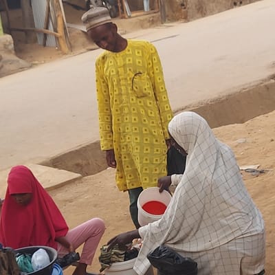 The image displays an outdoor scene with several individuals and simple structures, suggesting a community or village setting. The ground appears to be made of dirt or sand.
In the foreground and center-left, a man stands wearing a light-colored cap (possibly kufi) and a long-sleeved yellow tunic or caftan. The tunic has a repeating pattern of small, darker yellow squares or diamonds. His face is partially visible, and he appears to be looking towards the right side of the frame.
To the right of the man, another individual is partially visible, mostly obscured by a white, checkered garment that resembles a long robe or hijab. This person is holding a white bucket with a bright red interior, which suggests it might contain water or another liquid. Their face is not clearly visible.
Further to the right and lower in the frame, a young child is visible, wearing a pink or reddish garment that covers their head and most of their body. Only a portion of their face is seen, looking down or towards the front. Another white bucket, possibly with clothes or other items inside, is near this child.
In the background, there are indications of simple structures, possibly walls made of mud or concrete, and some wooden or metal elements suggesting fences or basic shelter. The overall lighting suggests it is daytime, possibly bright and sunny, given the lack of deep shadows. There is no visible text in the image.