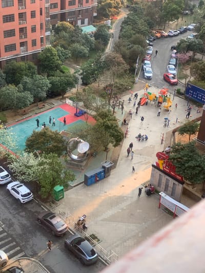 The image captures an aerial view of a residential area in Wuhan, China, located at coordinates 30.502 latitude and 114.476 longitude. The scene features a paved area with a basketball court, a playground, and a parking lot.
A group of people are seen playing basketball on the court, while others are gathered around the playground, which includes a slide, a climbing structure, and a swing set. Children are playing on the playground, while adults supervise or socialize nearby. Several cars are parked in the lot, with a few vehicles visible driving on the street.
The environment is characterized by a mixture of buildings and greenery. Several trees surround the paved area, with the leaves in various shades of green and brown. Buildings line the street, with one building directly behind the photographer.