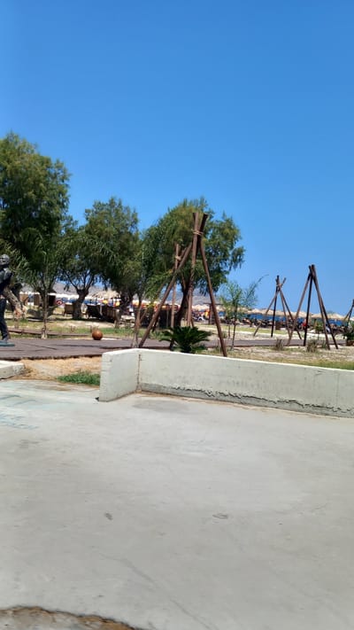 The image depicts a sunny day in Tigaki, Greece, with a clear blue sky dominating the upper portion of the frame. In the foreground, a concrete path or walkway is visible, with a textured, off-white retaining wall to the left. Beyond the wall, a wooden boardwalk leads to a park-like area with scattered trees and vegetation. Several wooden A-frame structures, possibly for swings or decorative purposes, are positioned in the mid-ground.

In the background, a beach scene unfolds. Rows of thatched umbrellas and lounge chairs are set up on the sand, indicating a recreational area. The sea is visible in the distance, merging with the horizon under the bright sky.

On the left side of the boardwalk, a dark-colored statue or sculpture of a person is partially visible. A round, brown object, possibly a ball, rests on the boardwalk near the statue. Some people are scattered among the beach chairs, but they are not in focus. The overall scene suggests a relaxed, coastal atmosphere typical of a Greek beach resort.