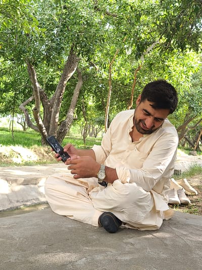 A man is sitting on the ground in an outdoor setting, holding a smartphone. He is wearing a light-colored traditional Pakistani outfit, including a shalwar and kameez. He has a beard and short, dark hair. He appears to be looking at the phone with a smile, suggesting he is engaged in a pleasant activity, possibly communicating or browsing.

The scene appears to be a park or garden area with numerous trees and green foliage. The ground is a mix of concrete and dirt, with some grassy areas visible. The lighting suggests it is daytime, likely during the morning or afternoon, with sunlight filtering through the leaves. The overall atmosphere seems relaxed and casual.

Notable details include the man's wristwatch, which has a silver band and a round face. The smartphone he is holding is black. The trees in the background are bare in some parts, with thick foliage in others, indicating a possibly temperate climate or a specific season. The orientation of the image is rotated, with the man appearing to be sitting upright and the background falling away to the side.

The location context is Kan Mehtarzai, Pakistan, which provides a geographical reference for the scene. There is no visible text in the image.