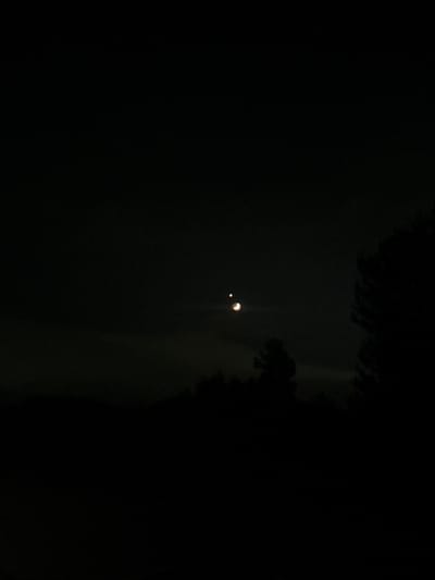 The image depicts a nighttime scene over Kraishte, Bulgaria, featuring celestial bodies and silhouetted natural elements. A crescent moon, illuminated from the right, is visible in the dark sky. To its left, a bright point of light, likely a planet or star, is also present. The foreground is dominated by the dark silhouettes of trees and possibly hills, suggesting a rural or natural setting within or near the city. The overall atmosphere is calm and serene, characteristic of a clear night.