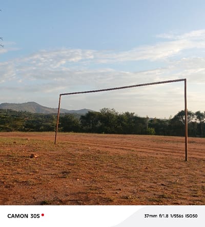 A rusty metal goalpost stands on a dirt field, with sparse green vegetation peeking through the red earth. In the background, a line of trees and a gently sloped hill meet a sky filled with soft, scattered clouds. The light suggests either sunrise or sunset, casting a warm glow over the scene. The field looks arid and untouched.