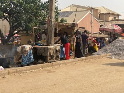 This image depicts an active outdoor market scene situated along an unpaved road in Makera, Nigeria. The foreground presents a light brown dirt surface transitioning to a concrete curb.

The midground is occupied by multiple makeshift market stalls constructed from poles and covered with tarps or various fabrics. A prominent concrete utility pole stands centrally. Approximately fifteen individuals are visible throughout the market area. On the far left, a person in a plaid shirt and patterned skirt is bent over a low cooking apparatus, from which smoke rises. Near the center, individuals are seen standing and interacting; one woman wears a black abaya with a head covering, and another, in a black top and red trousers, leans into a stall. To the right, a group of approximately five individuals sits on the ground adjacent to a substantial pile of grey gravel or crushed stone. A red and white striped market umbrella is partially visible over a stall further to the right.

The background reveals residential buildings with various roofing materials, predominantly corrugated metal. Several roofs feature installed solar panels. A solar-powered street light pole is also visible among the structures. The sky overhead is clear.