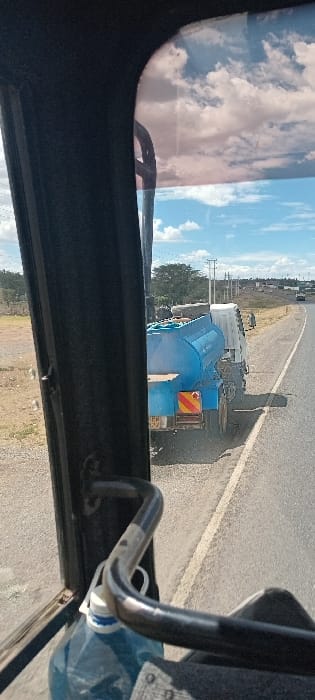 From within a vehicle, a bright day reveals a flat, slightly grassy landscape. An asphalt road stretches into the distance, marked by a white line. On the road, a truck with a blue tank and trailer is visible. Utility poles stand along the road. Reflections of clouds and sky are seen on the vehicle's windows. A blue water bottle is positioned inside the vehicle.