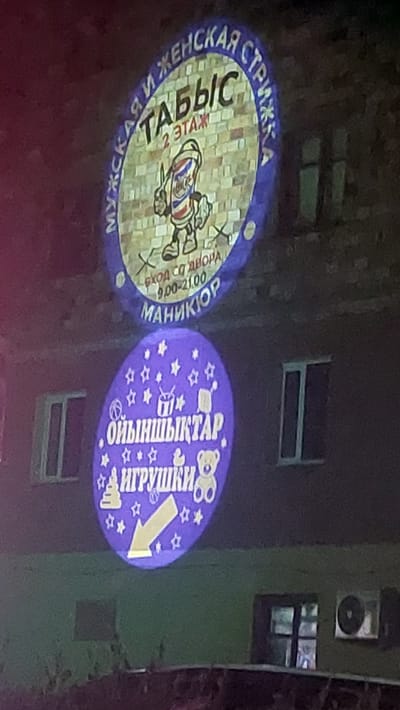 This media file captures a night scene in Shahtinsk, Kazakhstan, featuring two projected signs on the side of a building. The building appears to be dark, with windows visible. The lighting suggests it's nighttime, with the projections serving as illumination and advertisements.
The sign on the left is oval-shaped and primarily displays text and an illustration related to a barbershop. It features Russian text that translates to "Women's Haircut," and below that, "Tabiс." There's also information about "Entrance from the courtyard" and operating hours from "9:00-21:00," along with "Manicure." The illustration depicts a cartoon barber with a red, white, and blue striped pole. The text "Men's Haircut" is also visible around the bottom edge of this sign.
The sign on the right is also oval-shaped and is predominantly purple with white stars and graphics. The Russian text clearly reads "Oyinshyqtar" (Toys) and "IGRYSHKI" (Toys). This sign is decorated with cartoon teddy bears, stars, and a prominent yellow arrow pointing downwards and to the right, indicating direction.
The overall scene is lit by the projected images, creating a somewhat surreal and atmospheric effect against the dark building. The content of the signs suggests these are advertisements for local businesses: a barbershop and a toy store, possibly located within the building or nearby. The focus is on the brightly lit projections, with the background building and its architectural features serving as a backdrop.