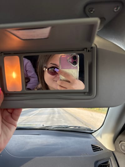 The image is taken from inside a car, looking towards the passenger side vanity mirror which is open. The mirror reflects a young woman taking a selfie. She is wearing sunglasses and has purple nail polish. She is holding a phone with a sparkly pink case and a diamond-studded border. A light is illuminated on the left side of the vanity mirror. Outside the car, a road is visible through the windshield, suggesting the car is in motion on a street or highway. The setting is likely a public road in or near Λούρος, Greece, based on the provided location hint.
