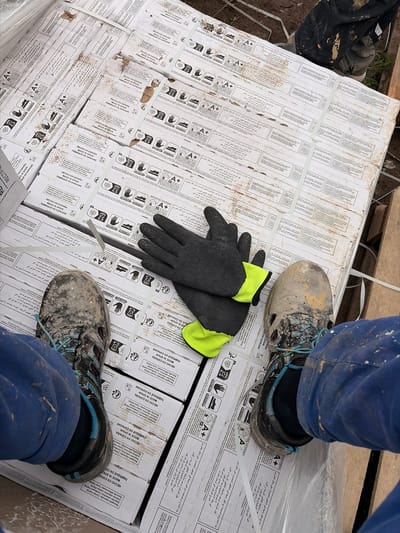 In the city of Příbram, Czechia, a close-up shot captures a construction worker's perspective, looking down at a stack of white cardboard boxes. The boxes are marked with multilingual text, including Arabic, Spanish, and French, and display various symbols indicating product information and handling instructions. A pair of black work gloves with neon yellow cuffs rests on top of the boxes. The worker's muddy work boots and blue work pants are visible in the foreground, suggesting an outdoor construction site. The ground appears to be dirt and debris, with some wooden planks and wire fencing visible in the background. The lighting suggests it is daytime.