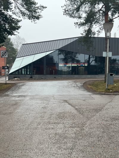 The image depicts the Aalto University Metro Station in Espoo, Finland, on an overcast day. The central subject is the modern building, characterized by an angular design featuring a dark, possibly metallic, segmented roof and a complex glass facade. The glass sections are a mix of transparent panels, reflecting the surrounding trees and the grey sky, and opaque white panels that create sharp, geometric lines. Visible through the transparent glass are interior lights and signs, one of which displays "Aalto-yliopisto" and a metro "M" logo.

In the foreground, a coarse, grey paved or asphalt surface, appearing damp and exhibiting tire marks, forks into two paths. To the left, a portion of a red-brick building is visible, alongside a circular white sign with a diagonal red line, indicating "no stopping" or "no parking," and a directional sign with an arrow pointing left and text that includes "Maanmittauslaitos." To the right, a utility pole with a unique concrete structure is present; a tree trunk appears to grow through this structure. A grey trash receptacle is mounted on this pole. Sparse, bare-branched trees, primarily pines, frame the scene from the left, right, and background. The sky is uniformly grey and overcast. No individuals are visible in the scene, and no explicit actions or interactions are depicted.