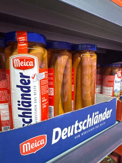 The image shows a shelf in a retail store, likely a supermarket, in Waldshut, Germany. On the shelf are several jars of "Meica" brand sausages, specifically labeled "Schlänländer". The jars are transparent, revealing the cooked sausages inside, packed in liquid. The branding on the jars and the prominent blue display box below the shelf clearly indicate the product. The blue box has the text "deutschländer vom würstchen das beste" (German sausages, the best sausages) printed in white lettering, with a red "Meica" logo on the left. The overall environment suggests a typical grocery store setting with product displays. There are no people visible in the image.