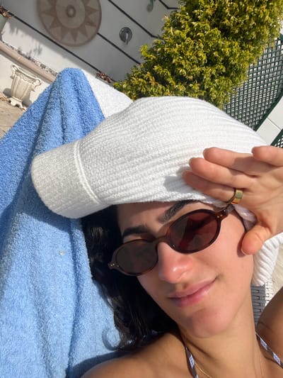 This close-up, high-angle image depicts a person relaxing outdoors on a bright, sunny day, likely in Èze, France. The subject is lying down, partially covered by a white, textured hooded garment or towel, which frames their head. Their face is partially visible, with dark-framed, round sunglasses covering their eyes. A gold-toned band ring is worn on a finger of their right hand, which is resting near their temple. A blue terry cloth towel is draped beneath them, suggesting a poolside or lounge setting. A strap from a striped bikini top is visible on their shoulder.

The background reveals an outdoor environment with lush green foliage from a dense bush, situated next to a dark green lattice or trellis structure. A white wall with vertical paneling is also visible, adorned with a circular, light-colored medallion featuring a starburst design and a small, white decorative urn. The strong sunlight casts clear shadows, indicating a bright daytime setting, possibly midday or early afternoon. The overall atmosphere conveys relaxation and tranquility in a well-maintained outdoor space. There is no visible text in the image.