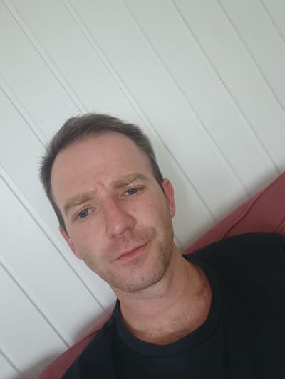 A man is pictured indoors, likely in an apartment, as indicated by the wood-paneled wall. He is wearing a black t-shirt and is leaning against a red cushion, possibly from a sofa. The lighting suggests a natural light source, casting soft shadows. The man's expression is neutral. The image does not provide any specific visual cues to pinpoint a location within Drammen, Norway.