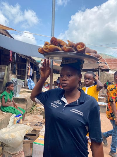 This image captures a street market scene in Igbara, Nigeria. In the foreground, a woman balances a large metal tray filled with what appears to be dried meat or animal skin on her head. She is wearing a navy blue polo shirt with white trim and text that reads "QUEENSFERRY CROSSING" and "Official Opening 04 September 2017". She is looking to her left, with her right hand raised in the air, possibly to steady the tray or to interact with someone off-camera.

Behind her, a young boy in a yellow shirt is visible, also looking to his left. To the left of the woman, a young girl in a green dress is seated on concrete steps. Several market stalls and structures made of wood and corrugated metal are present in the background, along with various goods such as bags, bottles, and baskets. The ground appears to be unpaved earth. The sky is partly cloudy.
