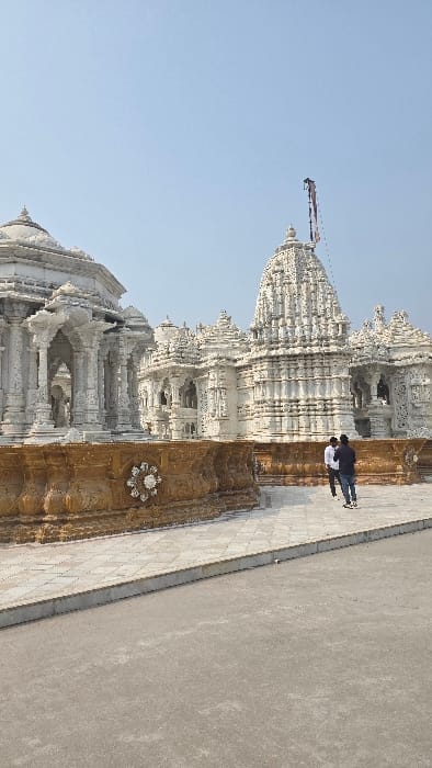 A tall, ornate white temple dominates the frame against a clear blue sky. Two figures stand near its base, possibly admiring the structure. The temple's design includes intricate carvings and multiple levels, giving it a tiered appearance. A brownish-gold wall extends along the right side, adding contrast to the white architecture. The surrounding area appears paved, suggesting a well-maintained site.