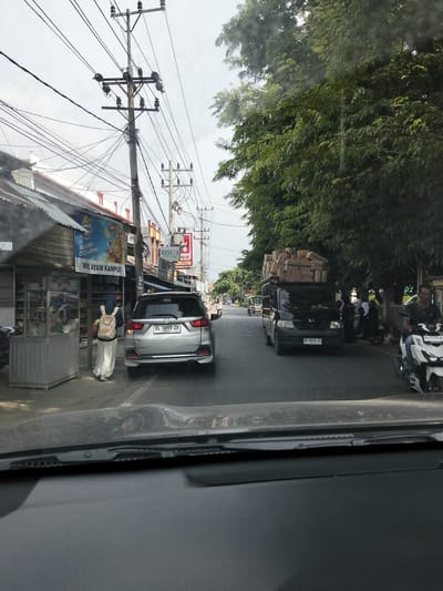 This media file captures a street scene in Keudematangglumpangdua, Indonesia. The viewpoint is from inside a vehicle, looking forward. On the left side of the road, there is a small food stall with a sign advertising "MIE AYAM KAMPUS" (Campus Chicken Noodles). A person wearing a blue headscarf and a beige backpack is standing near the stall. Further down the road, a silver SUV is parked.
Across the street, on the right, dense green trees line the roadside. Several people are gathered, some with motorcycles. A black truck overloaded with boxes is moving down the street, followed by a white scooter. The weather appears to be overcast, suggesting it might be a cloudy day. The overall scene depicts a typical street view with everyday activities of people and vehicles in an Indonesian town. Visible text includes brand names, store advertisements, and license plate numbers. The time is indicated as 12:29 on the silver SUV's license plate.