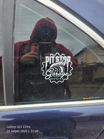 The image displays a close-up, angled view of the rear passenger door and window of a dark blue vehicle, located in Dolna Beshovitsa, Bulgaria. A white vinyl sticker, featuring a gear outline, is affixed to the exterior of the window. The sticker design incorporates the text "PIT STOP" above a turbocharger illustration, with "Garage REPAIRS" positioned beneath it. The window glass is visibly dirty, covered in dust and specks. A faint "3M TINT" logo is present in the lower right corner of the window.
A person, wearing a dark red or maroon hooded sweatshirt, is reflected in the car window. This individual is holding a dark-colored smartphone in their right hand, aimed toward the window, and a separate object with a red handle in their left hand. Their face is obscured by the reflection.
Through the window's reflection, an outdoor background is discernible, including a portion of a light-colored building and dark, bare vegetation under an overcast sky. The image was recorded by a Galaxy S25 Ultra on March 24, 2026, at 10:48.