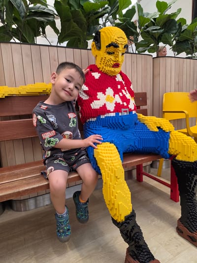 A young boy is sitting on a wooden bench next to a large Lego sculpture of a man. The boy is wearing a grey t-shirt with Marvel characters and grey shorts, and blue sandals. He is smiling and leaning his head on the Lego man's arm. The Lego man is wearing a red shirt with a floral pattern and blue pants. He is sitting with his legs extended. In the background, there are tall green plants and a wooden wall. A yellow chair is visible to the right. The image appears to be taken indoors. The location is not Mladá Boleslav, Czechia.